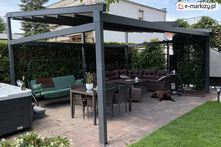 Zdjęcie prezentuje otwartą, wolnostojącą pergolę Square MOL o konstrukcji aluminiowej w kolorze antracytowym, zlokalizowaną na przydomowym tarasie. System zadaszenia składa się z dwóch niezależnych modułów dachowych, które są obecnie rozwinięte, odsłaniając poprzeczne belki konstrukcyjne wspierające mechanizm i zapewniając cień na tarasie. Pod pergolą zaaranżowano przestronną strefę rekreacyjną: po lewej stronie widoczne jest nowoczesne jacuzzi z ciemną obudową i białą niecką, obok którego znajduje się huśtawka ogrodowa z zielonym materacem. W centralnej części ustawiono duży stół jadalniany z ciemnego technorattanu z szklanym blatem i dopasowanymi krzesłami, a w głębi znajduje się narożna sofa z licznymi poduszkami, przy której odpoczywa duży, czekoladowy pies. Na tarasie z wielkoformatowych płyt kamiennych stoi również telewizor na mobilnym stojaku. Całość otacza starannie wypielęgnowana roślinność, w tym wysoki żywopłot z tui oraz pnącza na pergoli po prawej stronie. W tle widoczny jest fragment jasnej elewacji sąsiedniego budynku oraz zielony trawnik, co nadaje miejscu kameralny i luksusowy charakter.