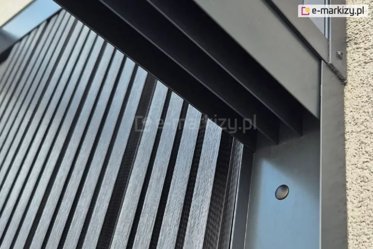 Górna prowadnica dla systemu szklanych szyb przesuwnych Miros zamocowana wewnątrz profilu doszczelniającego U przykręconego do ściany elewacji. W tle za prowadnicą widoczna ściana z dekoracyjnych lameli drewnianych w kolorze antracytowym, ułożonych pionowo.