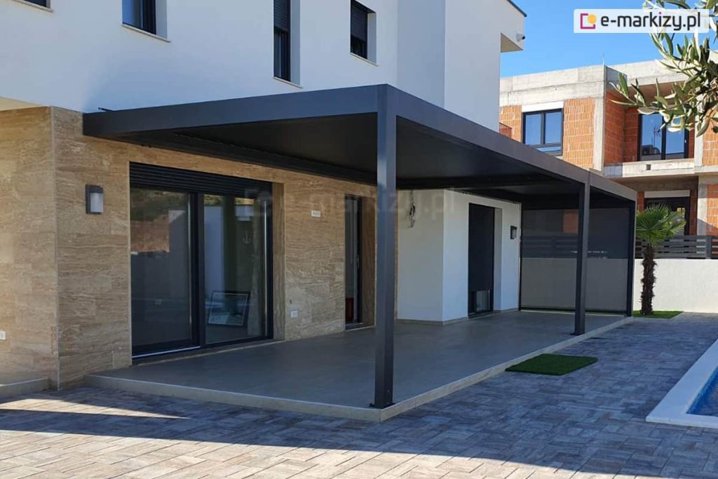 Pergola SB400 ustawiana na tarasie domu z jasną elewacją nad tarasem betonowym w pobliżu basenu oraz podwórka wyłożonego kostką