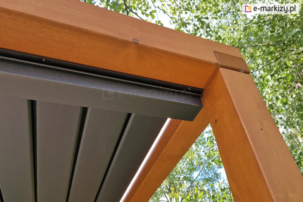 Zdjęcie prezentuje techniczne zbliżenie na detal montażowy narożnika pergoli drewnianej z zainstalowanym systemem rynnowym zadaszenia lamelowego SB400 MD. Solidna belka z drewna klejonego w kolorze jasnego miodu łączy się pod kątem prostym z pionowym słupem nośnym, a wzdłuż jej wewnętrznej krawędzi zamontowana jest aluminiowa rynna w kolorze antracytowym. Rynna znajduje się bezpośrednio pod płaszczyzną zamkniętych, ciemnoszarych lameli, co zapewnia skuteczne przejmowanie wody opadowej z dachu. Elementy stalowe i aluminiowe są precyzyjnie dopasowane do drewnianej ramy, a w górnej części rynny widoczne są drobne śruby montażowe. W tle, ponad konstrukcją, widoczne są gęste korony drzew liściastych z zielonymi liśćmi oraz jasne, lekko zachmurzone niebo.