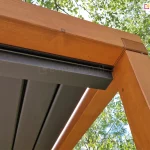 Zdjęcie prezentuje techniczne zbliżenie na detal montażowy narożnika pergoli drewnianej z zainstalowanym systemem rynnowym zadaszenia lamelowego SB400 MD. Solidna belka z drewna klejonego w kolorze jasnego miodu łączy się pod kątem prostym z pionowym słupem nośnym, a wzdłuż jej wewnętrznej krawędzi zamontowana jest aluminiowa rynna w kolorze antracytowym. Rynna znajduje się bezpośrednio pod płaszczyzną zamkniętych, ciemnoszarych lameli, co zapewnia skuteczne przejmowanie wody opadowej z dachu. Elementy stalowe i aluminiowe są precyzyjnie dopasowane do drewnianej ramy, a w górnej części rynny widoczne są drobne śruby montażowe. W tle, ponad konstrukcją, widoczne są gęste korony drzew liściastych z zielonymi liśćmi oraz jasne, lekko zachmurzone niebo.