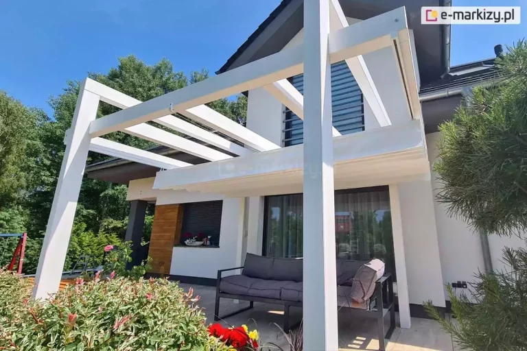 Pergola drewniana Moza z dachem rzymskim Supro zwiniętym na jedną stronę, dzięki czemu krokwie dachowe zostały odsłonięte i widoczny jest system sterowania ręcznego sznurkowego. pod dachem ustawiono meble tarasowe z szarymi poduchami. Na pierwszym tle kolorowe rośliny ogrodowe, a nad pergolą niebieskie bezchmurne niebo.