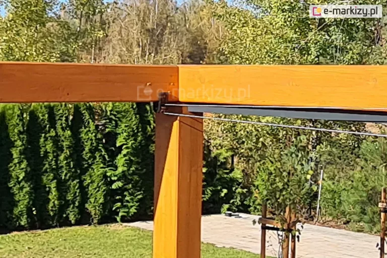 Konstrukcja drewniana nad taras koro z rozwijanym zadaszeniem tkaninowym supro elektrycznym, którego elementy przesuwane są za pomocą linek stalowych. W tle bogata, zielona roślinność ogrodu oraz wczesnowiosenne drzewa pozbawione jeszcze liści.