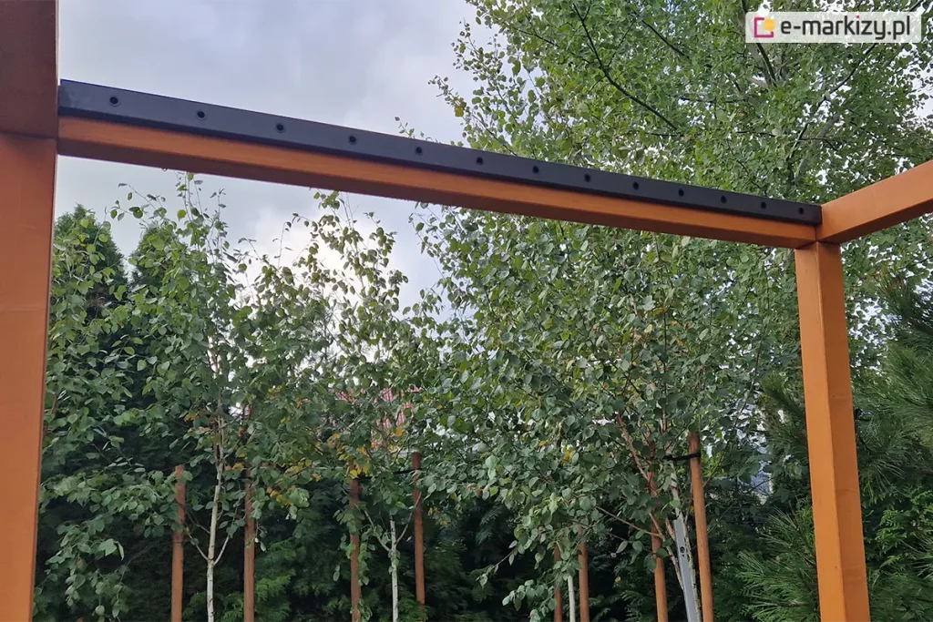 Zdjęcie przedstawia perspektywę z dołu na narożnik drewnianej konstrukcji pergoli w trakcie prac montażowych systemu dachu lamelowego SB400 MD. Rama wykonana z belek z drewna klejonego o ciepłym, pomarańczowo-brązowym odcieniu została wyposażona w czarne, aluminiowe listwy boczne. Listwy te, przytwierdzone do górnej części belek, posiadają seryjnie rozmieszczone, okrągłe otwory montażowe, w których docelowo osadzone zostaną łożyska i trzpienie obrotowe ruchomych lameli. Powierzchnia drewna jest wilgotna, z widocznymi kroplami wody, co podkreśla surowe warunki montażowe. W tle, ponad linią belek, widoczne są gęste korony drzew liściastych z zielonymi liśćmi, prześwitujące błękitne niebo oraz fragmenty sąsiedniej zabudowy jednorodzinnej z ciemnymi dachami.