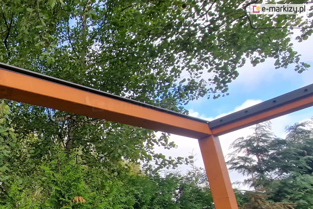 perspektywę z dołu na narożną część drewnianej konstrukcji pergoli podczas montażu systemu dachu lamelowego SB400 MD. Solidne belki z drewna klejonego o ciepłym, pomarańczowo-brązowym odcieniu są połączone pod kątem prostym, tworząc ramę nośną zadaszenia. Na górnej powierzchni belek zamontowano czarne, aluminiowe listwy montażowe, które są przygotowane do osadzenia w nich ruchomych piór (lameli). Listwy posiadają fabryczne otwory montażowe rozmieszczone w regularnych odstępach. Na powierzchni drewna widoczne są ślady wilgoci i pojedyncze krople wody. Tło zdjęcia stanowi gęsta korona drzewa liściastego o zielonych liściach, a w prawym dolnym rogu widoczny jest fragment ogrodu z iglakami pod jasnym, lekko zachmurzonym niebem.