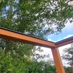 perspektywę z dołu na narożną część drewnianej konstrukcji pergoli podczas montażu systemu dachu lamelowego SB400 MD. Solidne belki z drewna klejonego o ciepłym, pomarańczowo-brązowym odcieniu są połączone pod kątem prostym, tworząc ramę nośną zadaszenia. Na górnej powierzchni belek zamontowano czarne, aluminiowe listwy montażowe, które są przygotowane do osadzenia w nich ruchomych piór (lameli). Listwy posiadają fabryczne otwory montażowe rozmieszczone w regularnych odstępach. Na powierzchni drewna widoczne są ślady wilgoci i pojedyncze krople wody. Tło zdjęcia stanowi gęsta korona drzewa liściastego o zielonych liściach, a w prawym dolnym rogu widoczny jest fragment ogrodu z iglakami pod jasnym, lekko zachmurzonym niebem.