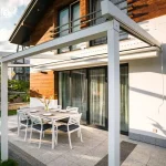 Biała aluminiowa pergola tarasowa Simple Tarasola o lekkiej konstrukcji przyściennej, zamontowana nad tarasem wyłożonym jasnoszarymi płytami. Stelaż składa się z poprzecznej belki frontowej wspartej na dwóch pionowych słupach o kwadratowym przekroju oraz prowadnic dachowych biegnących od elewacji budynku. Tkanina zadaszenia jest w całości zwinięta, co odsłania konstrukcję i pozwala na pełne nasłonecznienie strefy wypoczynkowej. Pod pergolą ustawiono zestaw obiadowy składający się z prostokątnego stołu z szarym blatem oraz sześciu białych krzeseł z grafitowymi siedziskami. Elewacja domu łączy biały tynk z panelami o strukturze drewna, a szerokie przeszklone drzwi prowadzą bezpośrednio na taras. Na jasnej ścianie widoczna jest zawieszona biała korba do manualnej obsługi dachu oraz wyraźne cienie rzucane przez konstrukcję w świetle dziennym.