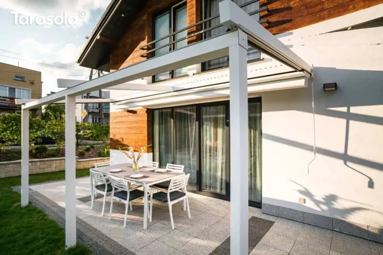 Biała aluminiowa pergola tarasowa Simple Tarasola o lekkiej konstrukcji przyściennej, zamontowana nad tarasem wyłożonym jasnoszarymi płytami. Stelaż składa się z poprzecznej belki frontowej wspartej na dwóch pionowych słupach o kwadratowym przekroju oraz prowadnic dachowych biegnących od elewacji budynku. Tkanina zadaszenia jest w całości zwinięta, co odsłania konstrukcję i pozwala na pełne nasłonecznienie strefy wypoczynkowej. Pod pergolą ustawiono zestaw obiadowy składający się z prostokątnego stołu z szarym blatem oraz sześciu białych krzeseł z grafitowymi siedziskami. Elewacja domu łączy biały tynk z panelami o strukturze drewna, a szerokie przeszklone drzwi prowadzą bezpośrednio na taras. Na jasnej ścianie widoczna jest zawieszona biała korba do manualnej obsługi dachu oraz wyraźne cienie rzucane przez konstrukcję w świetle dziennym.
