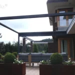 Zdjęcie przedstawia widok z boku na przyścienną pergolę tarasową Tarasola Flat o konstrukcji aluminiowej w kolorze antracytowym, której dach tkaninowy w kolorze jasnokremowym jest niemal całkowicie zwinięty w stronę elewacji budynku. Tkanina dachu spoczywa w formie ciasno złożonego pakietu pod dedykowanym daszkiem ochronnym, co odsłania pełną wysokość ramy i konstrukcję dachu płaskiego. Pergola przylega do elewacji budynku wykonanej z ciemnej cegły klinkierowej, z widocznym narożnym przeszkleniem i jasnym tynkiem na wyższej kondygnacji. Pod zadaszeniem znajduje się strefa wypoczynkowa z nowoczesną sofą ogrodową o szarej plecionej strukturze, ustawioną na tarasie wyłożonym jasnymi płytkami. Aranżację uzupełniają duże, podłużne donice ze stali corten o rdzawym odcieniu, w których rosną formowane w kule krzewy oraz białe kwiaty. W tle widoczny jest gęsty ogród z wysokimi drzewami iglastymi i liściastymi pod zachmurzonym niebem.