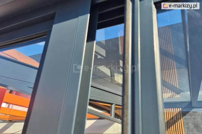 Zdjęcie prezentuje szczegółowy widok techniczny konstrukcji ramowej systemu przesuwnego Sotro, wykonanego z profili aluminiowych w kolorze antracytowym. Fotografia ukazuje sposób osadzenia pakietów szybowych w pionowych profilach oraz mechanizm wielotorowej prowadnicy, która umożliwia nachodzenie na siebie poszczególnych skrzydeł szklanych. Profile charakteryzują się smukłą linią i matowym wykończeniem, a widoczne uszczelki szczotkowe oraz systemowe wgłębienia zapewniają szczelność i płynność ruchu paneli. W odbiciu szyb oraz w prześwitach widoczne są elementy konstrukcyjne zadaszenia, w tym poziome lamele dekoracyjne w kolorze naturalnego drewna oraz fragmenty elewacji o strukturze tynku. Perspektywa z dołu podkreśla precyzję dopasowania elementów ramy oraz solidność montażu systemów jezdnych. Tło stanowi błękitne niebo oraz fragmenty architektury ogrodowej, które przenikają przez transparentne tafle szkła, co akcentuje wysoką przezierność zastosowanego szklenia.