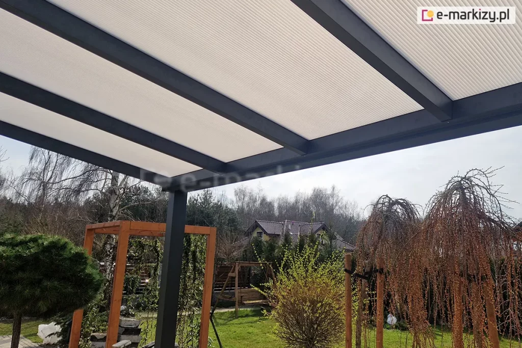 Widok od spodu na konstrukcję dachu zadaszenia tarasowego Modes, ukazujący połączenie materiałów i kolorystykę. Połacie dachowe wykonane są z poliwęglanu komorowego w kolorze mlecznym, który równomiernie rozprasza światło dzienne. Płyty są stabilnie osadzone na systemowych belkach aluminiowych malowanych proszkowo na kolor antracytowy, które tworzą wyraźny, geometryczny rytm. W tle, pod zadaszeniem, roztacza się widok na ogród z zielonym trawnikiem, ozdobnymi drzewami iglastymi oraz drewnianymi konstrukcjami ogrodowymi (pergola, huśtawka) na tle sąsiednich domów jednorodzinnych.