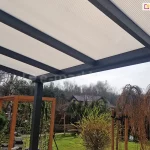 Widok od spodu na konstrukcję dachu zadaszenia tarasowego Modes, ukazujący połączenie materiałów i kolorystykę. Połacie dachowe wykonane są z poliwęglanu komorowego w kolorze mlecznym, który równomiernie rozprasza światło dzienne. Płyty są stabilnie osadzone na systemowych belkach aluminiowych malowanych proszkowo na kolor antracytowy, które tworzą wyraźny, geometryczny rytm. W tle, pod zadaszeniem, roztacza się widok na ogród z zielonym trawnikiem, ozdobnymi drzewami iglastymi oraz drewnianymi konstrukcjami ogrodowymi (pergola, huśtawka) na tle sąsiednich domów jednorodzinnych.