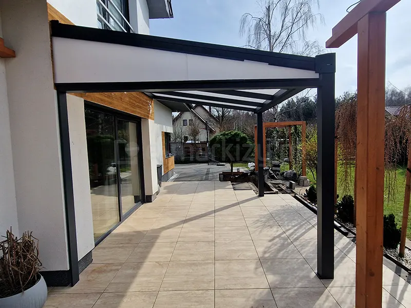 Zdjęcie prezentuje gotową realizację przyściennego zadaszenia tarasowego Modes, zamontowanego na nowoczesnym tarasie wyłożonym jasnobeżowymi płytami. Antracytowa konstrukcja aluminiowa wspiera dach wykonany z płyt poliwęglanu komorowego, a jej cechą charakterystyczną jest wypełnienie trójkątnego boku (tzw. klina) białą, nieprzezierną płytą, co zwiększa osłonę przed wiatrem i sąsiadami. Zadaszenie opiera się na dwóch solidnych słupach o przekroju kwadratowym, a cała struktura przylega do białej elewacji budynku z dekoracyjnym pasem o strukturze drewna. W tle widoczny jest ogród z trawnikiem, iglakami oraz fragmenty drewnianej pergoli i sąsiedniej zabudowy, co ukazuje wkomponowanie produktu w przestrzeń przydomową.