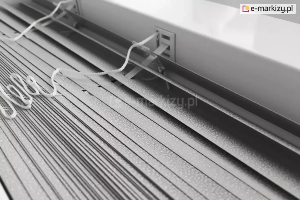 Zdjęcie prezentuje techniczne zbliżenie na żaluzje aluminiowe 16 mm w kolorze srebrnym lub jasnoszarym, ukazane z perspektywy górnej. Konstrukcja składa się z bardzo wąskich lameli o szerokości zaledwie szesnastu milimetrów, które charakteryzują się drobną, ziarnistą strukturą powierzchni typu "perforacja" lub "frost", odbijającą światło w sposób rozproszony. Lamele są ciasno ułożone jedna nad drugą, co podkreśla ich smukły profil, idealny do montażu w przestrzeniach z ograniczoną ilością miejsca, np. między listwami przyszybowymi. Centralnym punktem kadru jest miejsce wyjścia białej, plecionej drabinki sznurkowej z rynny górnej, która przechodzi przez precyzyjny, prostokątny otwór w metalowej obudowie. Białe sznurki operacyjne, służące do sterowania mechanizmem, są luźno ułożone na powierzchni zebranych lameli, prezentując swoją teksturę i splot. Tło stanowi fragment białej ramy okiennej lub ściany, co tworzy jasną i sterylną kompozycję kolorystyczną. Całość zdjęcia akcentuje lekkość, precyzję wykonania oraz minimalistyczny design, jaki oferuje żaluzja aluminiowa 16 mm w nowoczesnych systemach osłonowych.