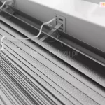 Zdjęcie prezentuje techniczne zbliżenie na żaluzje aluminiowe 16 mm w kolorze srebrnym lub jasnoszarym, ukazane z perspektywy górnej. Konstrukcja składa się z bardzo wąskich lameli o szerokości zaledwie szesnastu milimetrów, które charakteryzują się drobną, ziarnistą strukturą powierzchni typu "perforacja" lub "frost", odbijającą światło w sposób rozproszony. Lamele są ciasno ułożone jedna nad drugą, co podkreśla ich smukły profil, idealny do montażu w przestrzeniach z ograniczoną ilością miejsca, np. między listwami przyszybowymi. Centralnym punktem kadru jest miejsce wyjścia białej, plecionej drabinki sznurkowej z rynny górnej, która przechodzi przez precyzyjny, prostokątny otwór w metalowej obudowie. Białe sznurki operacyjne, służące do sterowania mechanizmem, są luźno ułożone na powierzchni zebranych lameli, prezentując swoją teksturę i splot. Tło stanowi fragment białej ramy okiennej lub ściany, co tworzy jasną i sterylną kompozycję kolorystyczną. Całość zdjęcia akcentuje lekkość, precyzję wykonania oraz minimalistyczny design, jaki oferuje żaluzja aluminiowa 16 mm w nowoczesnych systemach osłonowych.