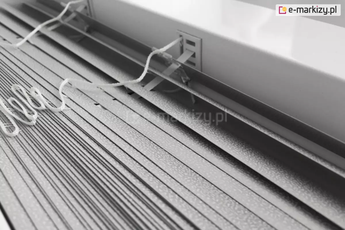 Zdjęcie prezentuje techniczne zbliżenie na żaluzje aluminiowe 16 mm w kolorze srebrnym lub jasnoszarym, ukazane z perspektywy górnej. Konstrukcja składa się z bardzo wąskich lameli o szerokości zaledwie szesnastu milimetrów, które charakteryzują się drobną, ziarnistą strukturą powierzchni typu "perforacja" lub "frost", odbijającą światło w sposób rozproszony. Lamele są ciasno ułożone jedna nad drugą, co podkreśla ich smukły profil, idealny do montażu w przestrzeniach z ograniczoną ilością miejsca, np. między listwami przyszybowymi. Centralnym punktem kadru jest miejsce wyjścia białej, plecionej drabinki sznurkowej z rynny górnej, która przechodzi przez precyzyjny, prostokątny otwór w metalowej obudowie. Białe sznurki operacyjne, służące do sterowania mechanizmem, są luźno ułożone na powierzchni zebranych lameli, prezentując swoją teksturę i splot. Tło stanowi fragment białej ramy okiennej lub ściany, co tworzy jasną i sterylną kompozycję kolorystyczną. Całość zdjęcia akcentuje lekkość, precyzję wykonania oraz minimalistyczny design, jaki oferuje żaluzja aluminiowa 16 mm w nowoczesnych systemach osłonowych.