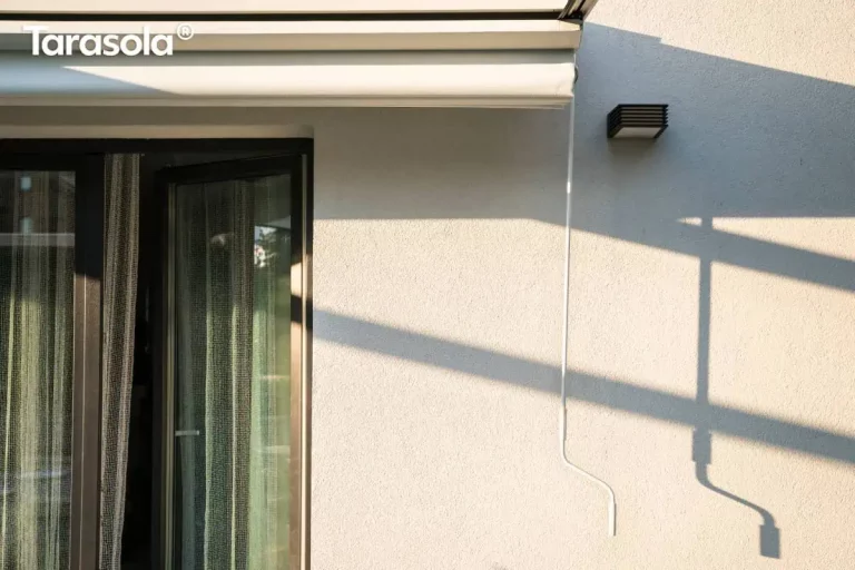 Zbliżenie na białą korbę ręczną służącą do sterowania zadaszeniem pergoli tarasowej Tarasola Simple. Metalowy drążek o ergonomicznym wygięciu w dolnej części zwisa pionowo, będąc połączonym z mechanizmem zwijającym tkaninę zamontowanym przy krawędzi profilu aluminiowego. Tło stanowi jasna, chropowata elewacja budynku z widocznym fragmentem ciemnych ram drzwi tarasowych oraz nowoczesną, czarną lampą elewacyjną o geometrycznym kształcie. Na ścianie rysują się wyraźne, ukośne cienie rzucane przez konstrukcję pergoli w świetle popołudniowego słońca.