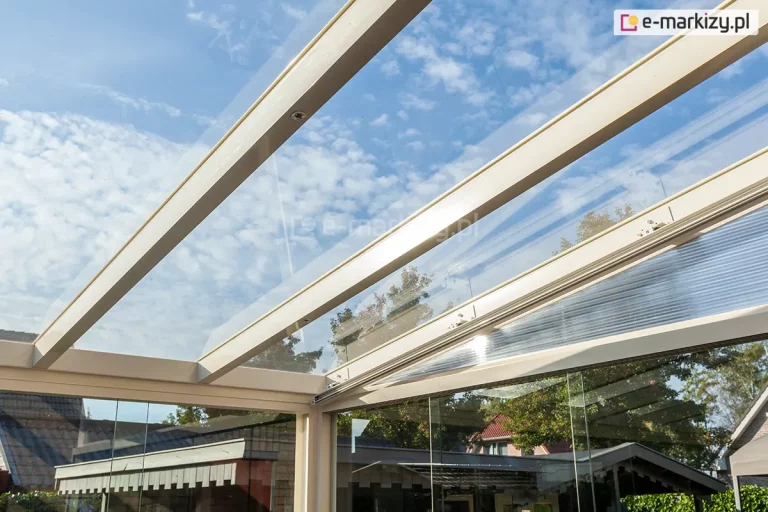 Zbliżenie na detale konstrukcyjne zadaszenia tarasowego Terano w rzadziej spotykanym, kremowym kolorze profili aluminiowych. Zdjęcie wykonane z perspektywy żabiej ukazuje transparentne panele szklane dachu, które są precyzyjnie osadzone na aluminiowych krokwiach. Konstrukcja dachu łączy się ze szklanymi ścianami przesuwnymi, tworząc szczelną i estetyczną zabudowę typu ogród zimowy. Przez przezroczyste połacie dachu widać błękitne niebo z delikatnym zachmurzeniem oraz wierzchołki drzew. W odbiciu szklanych tafli oraz w tle widoczne są sąsiednie zabudowania mieszkalne i ogrodowe.