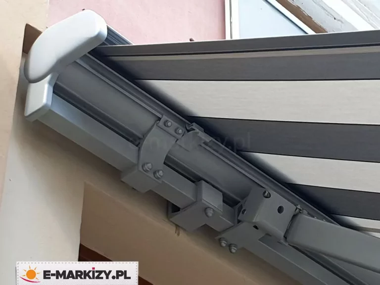 Techniczne zbliżenie na system montażowy oraz konstrukcję markizy tarasowej Palladio zamontowanej na jasnej elewacji budynku w niewielkiej wnęce. Kluczowym elementem widocznym na fotografii jest masywny, aluminiowy profil nośny o kwadratowym przekroju, polakierowany proszkowo na kolor srebrny. Do profilu tego, za pomocą solidnych stalowych uchwytów i śrub o wyraźnie zaznaczonych łbach, przytwierdzona jest kaseta markizy oraz mechanizm ramion przegubowych, a także uchwyt ścienny. Po prawej stronie kadru widoczny jest fragment przegubu ramienia, który odpowiada za stabilne rozkładanie poszycia. Sama tkanina markizowa charakteryzuje się nowoczesnym wzorem w szerokie, poziome pasy w kolorze ciemnoszarym i jasnoszarym (srebrnym), co nadaje całości minimalistyczny i techniczny wygląd. Widok od dołu pozwala na dokładną analizę punktów styku elementów mechanicznych oraz precyzję wykonania łączeń, które gwarantują wytrzymałość całej osłony przeciwsłonecznej.