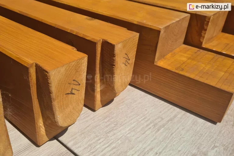 Zbliżenie zakończeń belek drewnianych ułożonych na płytkach gresowych. Dwie belki z widocznymi czopami z umieszczonych na nich cyframi 14 wykonanymi czarnym markerem. W tle dwie nacięte belki dachowe.