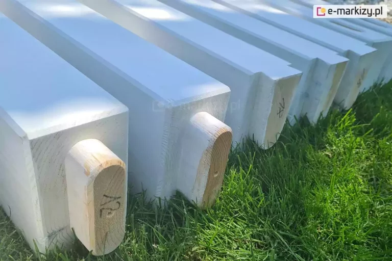 Zbliżenie elementów konstrukcji pergoli drewnianej Moza ułożonych na trawie, z widocznymi charakterystycznymi frezowaniami mocowania podpór i krokwi oraz naniesionymi na niemalowanych fragmentach oznaczeniami elementów w postaci cyfr, na pierwszym od lewej elemencie cyfra "12".
