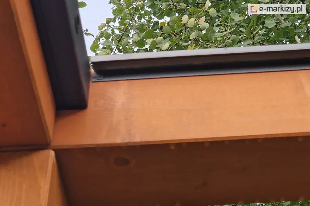 techniczny detal połączenia belek konstrukcyjnych pergoli drewnianej w narożniku, przygotowany pod montaż systemu lamelowego SB400 MD. Na miodowym, jasnobrązowym drewnie klejonym zamontowana jest czarna, aluminiowa listwa montażowa, która pełni jednocześnie funkcję okapową. Listwa jest przytwierdzona do górnej krawędzi poziomej belki za pomocą wkrętów, a jej profil jest precyzyjnie docięty i dopasowany do pionowego elementu ramy widocznego po lewej stronie. Na powierzchni drewna widoczne są ślady wilgoci w postaci kropel wody. W tle, ponad konstrukcją, rozpościera się widok na korony drzew liściastych z zielonymi i żółknącymi liśćmi pod jasnym niebem.