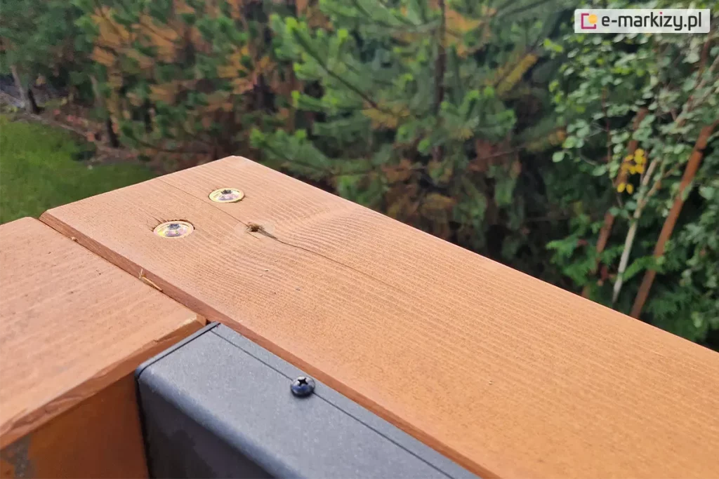 Zbliżenie połączenia narożnika belek konstrukcji drewnianej pergoli Koro z przymocowanym do jednej z belek profilem dachu lamelowego SB400 MD Selt. W tle widoczne rośliny iglaste.