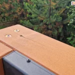 Zbliżenie połączenia narożnika belek konstrukcji drewnianej pergoli Koro z przymocowanym do jednej z belek profilem dachu lamelowego SB400 MD Selt. W tle widoczne rośliny iglaste.