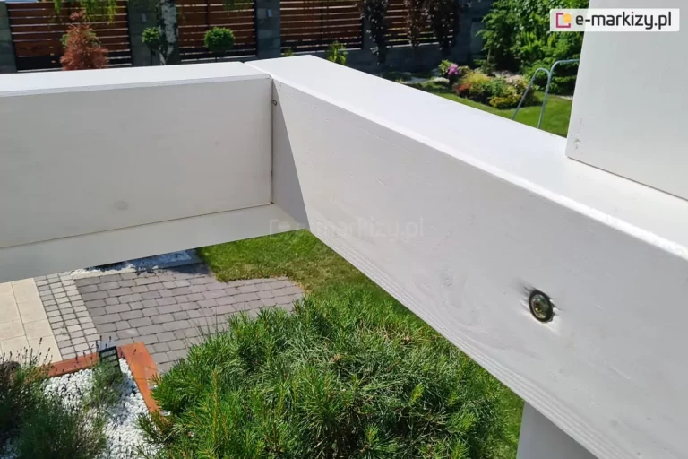Zbliżenie na narożnik pergoli drewnianej Moza z widoczną śrubą mocującą wieniec pergoli do jej bramki. W tle drzewko iglaste, fragment podwórka z kostki brukowej oraz płot z drewnianymi panelami.