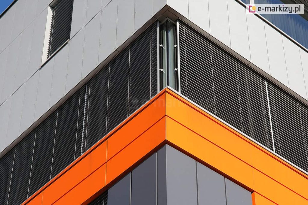 Narożnik nowoczesnego budynku biurowego z dużymi przeszkleniami wyposażonymi w zewnętrzne żaluzje fasadowe. System żaluzji obejmuje dwie przylegające do siebie fasady, z których każda osłonięta jest poziomymi lamelami aluminiowymi w kolorze antracytowym. Lamele są ustawione równolegle i prowadzone w prowadnicach bocznych mocowanych do ramy okna. Elewacja budynku wykończona jest panelami elewacyjnymi w dwóch kolorach: na dolnej części widoczne są panele w kolorze ciemnego grafitu oraz intensywnej pomarańczy, a górna część elewacji pokryta jest jasnoszarymi płytami kompozytowymi.