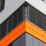 Narożnik nowoczesnego budynku biurowego z dużymi przeszkleniami wyposażonymi w zewnętrzne żaluzje fasadowe. System żaluzji obejmuje dwie przylegające do siebie fasady, z których każda osłonięta jest poziomymi lamelami aluminiowymi w kolorze antracytowym. Lamele są ustawione równolegle i prowadzone w prowadnicach bocznych mocowanych do ramy okna. Elewacja budynku wykończona jest panelami elewacyjnymi w dwóch kolorach: na dolnej części widoczne są panele w kolorze ciemnego grafitu oraz intensywnej pomarańczy, a górna część elewacji pokryta jest jasnoszarymi płytami kompozytowymi.