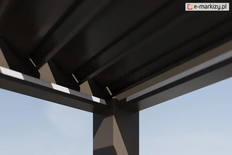 Zbliżenie na konstrukcyjny węzeł łączący pionową podporę z poziomymi belkami ramy i systemem ruchomego dachu w antracytowej pergoli lamelowej Marlo. Zdjęcie wykonane z perspektywy żabiej ukazuje detal osadzenia narożnego słupa o kwadratowym przekroju, na którym spoczywają dwie prostopadłe belki nośne ramy. Powyżej ramy widoczne są spodnie części aluminiowych lamel dachowych, które są ustawione w pozycji zamkniętej, tworząc szczelną i jednolitą płaszczyznę, pod nimi widoczny biały pas oświetlenia LED. Całość konstrukcji jest wykończona w matowym, antracytowym kolorze, a na krawędziach belek ramy widoczne są jasne refleksy świetlne podkreślające krawędzie profili. W tle, w szczelinach między elementami konstrukcyjnymi, widoczne jest pogodne, jasnobłękitne niebo.