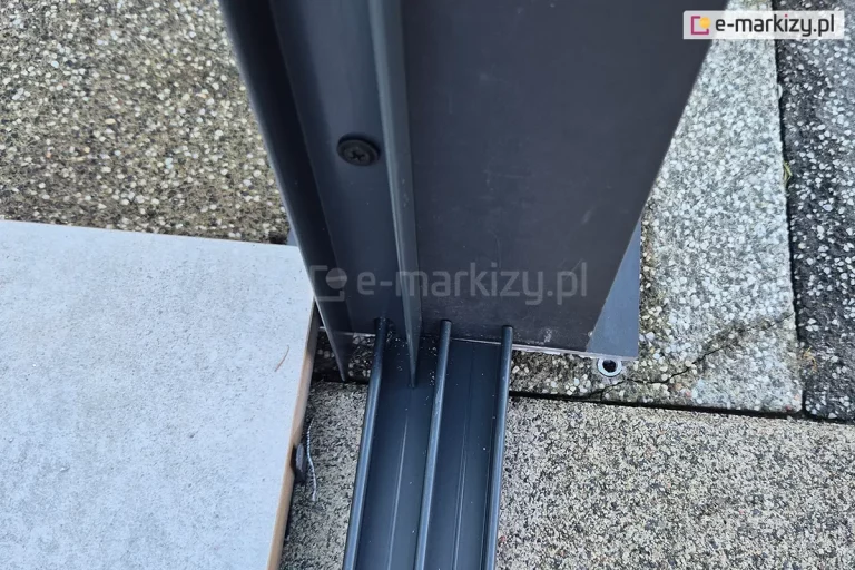 Zbliżenie na sposób połączenia prowadnicy dolnej do paneli szklanych Sotro oraz profilu bocznego doszczelniającego ścianę. Prowadnica mocowana do podłoża z betonowych płyt, a profil boczny U przykręcony do podpory zadaszenia.