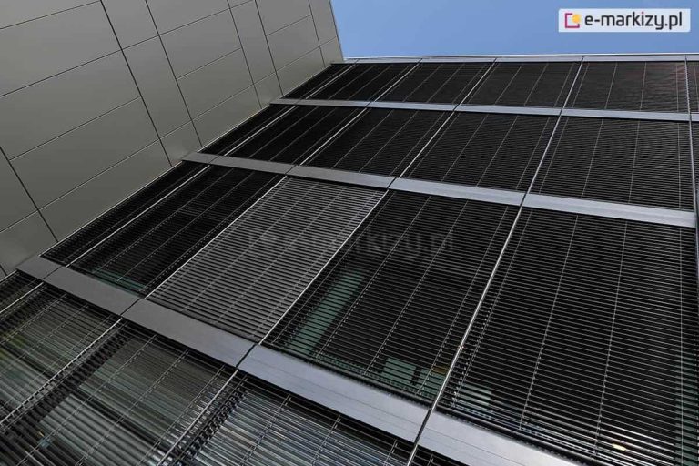 Fragment nowoczesnej elewacji budynku biurowego lub komercyjnego, sfotografowanej z ciekawej perspektywy, od dołu skierowanej ku górze. Elewacja wykonana jest z dużych, pionowych przeszklonych modułów osłoniętych zewnętrznymi żaluzjami fasadowymi C80 Oval łączonymi modułowo. Żaluzje składają się z gęsto rozmieszczonych, wąskich, poziomych aluminiowych lamel w kolorze ciemnoszarym lub grafitowym. Lamele prowadzone są w prowadnicach, wspólnych dla sąsiadujących ze sobą żaluzji. Całość fasady jest rytmicznie podzielona przez poziome pasy profili aluminiowych, oddzielające kolejne kondygnacje lub sekcje okienne. Po lewej stronie kadru widoczna jest sąsiadująca ściana pokryta gładkimi, jasnoszarymi panelami elewacyjnymi. Niebo w tle jest bezchmurne, co podkreśla kontrast pomiędzy jasnym otoczeniem a ciemną, zamkniętą żaluzją, która skutecznie ogranicza dostęp światła do wnętrza budynku.