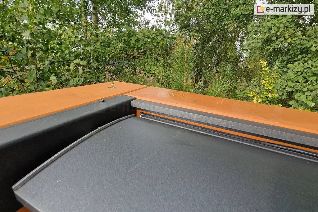 Narożnik drewnianej konstrukcji pergoli z zamocowanym do niej dachem lamelowym SB400 MD Selt z widocznym jednym lamelem w kolorze antracytowym. W tle widoczne liczne zielone drzewa i krzewy.