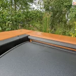 Narożnik drewnianej konstrukcji pergoli z zamocowanym do niej dachem lamelowym SB400 MD Selt z widocznym jednym lamelem w kolorze antracytowym. W tle widoczne liczne zielone drzewa i krzewy.