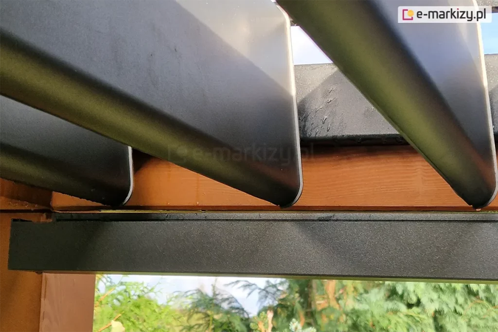 Zbliżenie rynny oraz zakończenia lamel dachowych w kolorze antracytowym zamocowanych na drewnianej konstrukcji. Rynny wzdłuż wysięgu odprowadzają wodę z lameli.