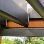 Zbliżenie rynny oraz zakończenia lamel dachowych w kolorze antracytowym zamocowanych na drewnianej konstrukcji. Rynny wzdłuż wysięgu odprowadzają wodę z lameli.