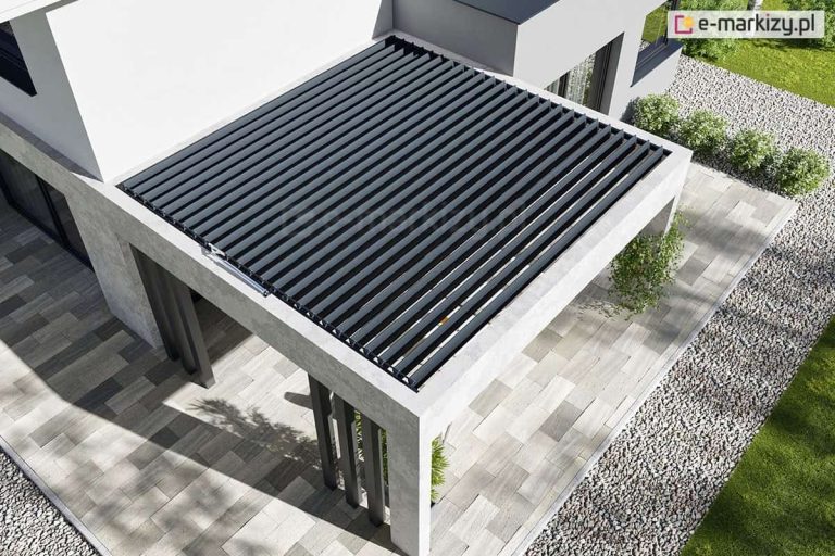 widok z góry na nowoczesną, betonową konstrukcję tarasu z zainstalowanym systemem dachu lamelowego Selt SB400 MD. Antracytowe, aluminiowe lamele są zamontowane wewnątrz jasnoszarej, betonowej ramy, która stanowi integralną część bryły budynku. Pióra dachu są lekko uchylone, zapewniając odpowiedni przepływ powietrza dla przestrzeni poniżej. Po lewej stronie, na krawędzi ramy, widoczny jest srebrny siłownik liniowy odpowiedzialny za automatyczne sterowanie kątem nachylenia lameli. Taras oraz ścieżki wokół domu wyłożone są wielkoformatowymi płytami brukowymi w różnych odcieniach szarości, a całość otacza pas jasnego żwiru dekoracyjnego oraz zadbany trawnik z rzędem niewielkich krzewów. Architektura budynku jest minimalistyczna, o białych i szarych elewacjach, co podkreśla techniczny i nowoczesny charakter zadaszenia.