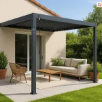 Nowoczesna pergola lamelowa Marlo w kolorze antracytowym, zamontowana w sposób przyścienny na jasnym tarasie. Konstrukcja zadaszenia wspiera się na dwóch pionowych słupach o kwadratowym przekroju, a jej tylna część jest przymocowana do beżowej elewacji budynku jednorodzinnego. Z lewej strony ramy widoczna jest czarna korba służąca do manualnej regulacji kąta nachylenia lameli dachowych, które są obecnie ustawione w pozycji zamkniętej. Pod pergolą wyznaczono strefę relaksu z beżową sofą ozdobioną zielonymi poduszkami, plecionym fotelem oraz niskim drewnianym stolikiem kawowym ustawionym na jasnym dywanie zewnętrznym. Nawierzchnia tarasu wykonana jest z dużych, jasnoszarych płyt betonowych, które graniczą z intensywnie zielonym, zadbanym trawnikiem. W terakotowej donicy po lewej stronie rośnie krzew ozdobny, a tło wypełnia gęsta ściana wysokich drzew i krzewów pod czystym, błękitnym niebem. Na elewacji domu widoczny jest wyraźny cień rzucany przez dach pergoli, co wskazuje na silne nasłonecznienie.