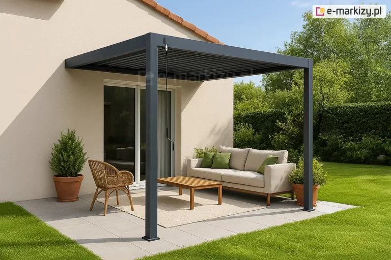 Nowoczesna pergola lamelowa Marlo w kolorze antracytowym, zamontowana w sposób przyścienny na jasnym tarasie. Konstrukcja zadaszenia wspiera się na dwóch pionowych słupach o kwadratowym przekroju, a jej tylna część jest przymocowana do beżowej elewacji budynku jednorodzinnego. Z lewej strony ramy widoczna jest czarna korba służąca do manualnej regulacji kąta nachylenia lameli dachowych, które są obecnie ustawione w pozycji zamkniętej. Pod pergolą wyznaczono strefę relaksu z beżową sofą ozdobioną zielonymi poduszkami, plecionym fotelem oraz niskim drewnianym stolikiem kawowym ustawionym na jasnym dywanie zewnętrznym. Nawierzchnia tarasu wykonana jest z dużych, jasnoszarych płyt betonowych, które graniczą z intensywnie zielonym, zadbanym trawnikiem. W terakotowej donicy po lewej stronie rośnie krzew ozdobny, a tło wypełnia gęsta ściana wysokich drzew i krzewów pod czystym, błękitnym niebem. Na elewacji domu widoczny jest wyraźny cień rzucany przez dach pergoli, co wskazuje na silne nasłonecznienie.