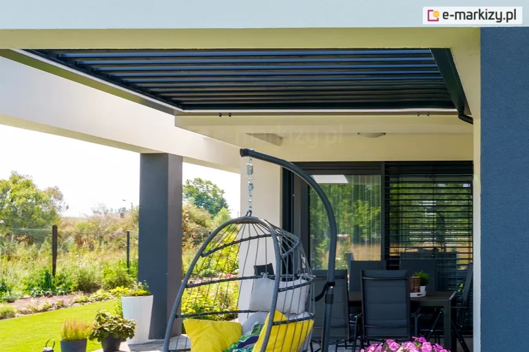 Zdjęcie przedstawia nowoczesne, bioklimatyczne zadaszenie lamelowe modelu SB400R, które zostało zintegrowane z istniejącą konstrukcją żelbetową tarasu. System składa się z ruchomych, aluminiowych piór w ciemnym kolorze antracytowym, które w tej konfiguracji są ustawione w pozycji zamkniętej, tworząc szczelną i płaską płaszczyznę sufitu. Konstrukcja dachu jest wpuszczona w białą wnękę stropu, co pozwala na zachowanie minimalistycznej estetyki i pełne ukrycie mechanizmów bocznych. Na wewnętrznej krawędzi obramowania widoczna jest zintegrowana listwa oświetleniowa LED, biegnąca wzdłuż boku zadaszenia. Taras jest urządzony w stylu nowoczesnym, z widocznym w centralnej części wiszącym fotelem typu kokon na czarnym stelażu z żółtymi poduszkami oraz zestawem mebli obiadowych z ciemnymi krzesłami w tle. Przeszklenia budynku są osłonięte zewnętrznymi żaluzjami fasadowymi w kolorze dopasowanym do lameli dachu. Całość znajduje się w otoczeniu zadbanej zieleni ogrodowej z trawnikiem i niskimi nasadzeniami roślinnymi.