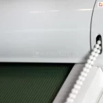 Zbliżenie kasety rolety LUX KOMBI z widocznym łańcuszkiem do sterowania żaluzją. Kaseta oraz prowadnice wykonane zostały w kolorze białym, współgrającym z ramą okna. Roletka została wyposażona w tkaninę o zielonym kolorze w delikatne pionowe paski.