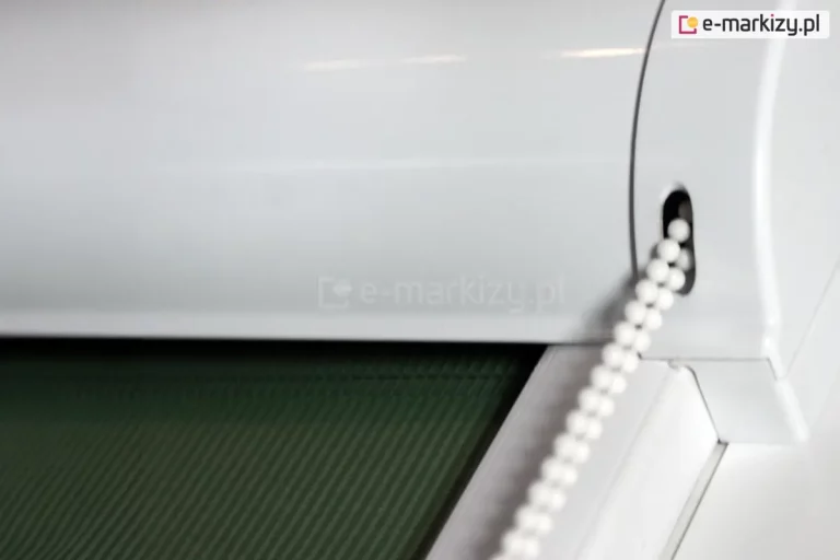 Zbliżenie kasety rolety LUX KOMBI z widocznym łańcuszkiem do sterowania żaluzją. Kaseta oraz prowadnice wykonane zostały w kolorze białym, współgrającym z ramą okna. Roletka została wyposażona w tkaninę o zielonym kolorze w delikatne pionowe paski.