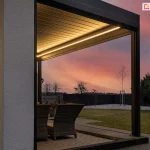 Pergola tarasowa SB 400 Aluprof z taśmowym oświetleniem LED o ciepłej barwie, ustawiona na tarasie z fotelami z rattanu z widocznym w tle różowym zachodem słońca