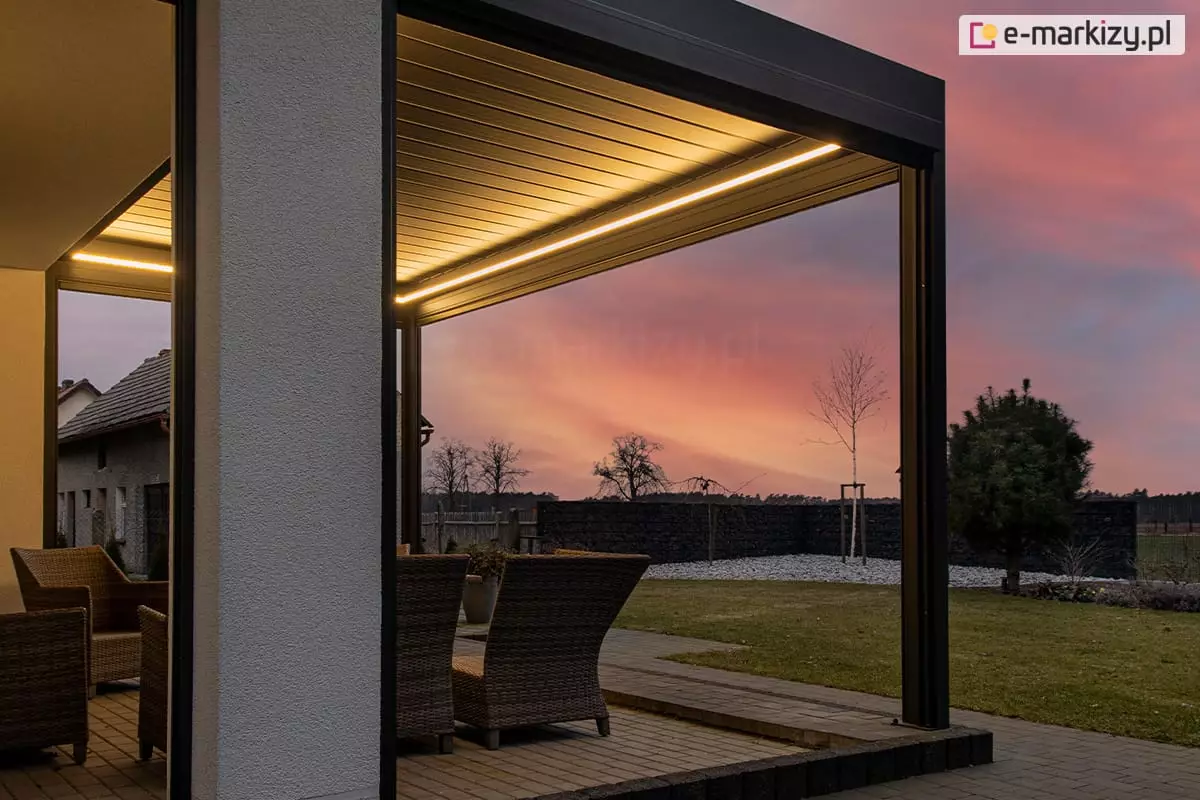Pergola tarasowa SB 400 Aluprof z taśmowym oświetleniem LED o ciepłej barwie, ustawiona na tarasie z fotelami z rattanu z widocznym w tle różowym zachodem słońca