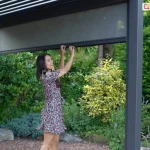 Pergola Gloria z napędem ręcznym korbowym do lamelowego dachu ustawiona w ogrodzie pełnym zielonych krzewów i drzew. Jedna ze ścian pergoli wyposażona została w roletę screen, która opuszcza w dół, ciągnąc dłońmi za profil kobieta o ciemnych włosach ubrana w sukienkę mini o kwiatowym wzorze. Pod pergolą widoczne oparcie krzesła w złoconym kolorze.