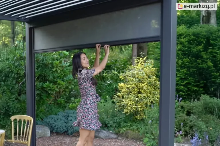 Pergola Gloria z napędem ręcznym korbowym do lamelowego dachu ustawiona w ogrodzie pełnym zielonych krzewów i drzew. Jedna ze ścian pergoli wyposażona została w roletę screen, która opuszcza w dół, ciągnąc dłońmi za profil kobieta o ciemnych włosach ubrana w sukienkę mini o kwiatowym wzorze. Pod pergolą widoczne oparcie krzesła w złoconym kolorze.