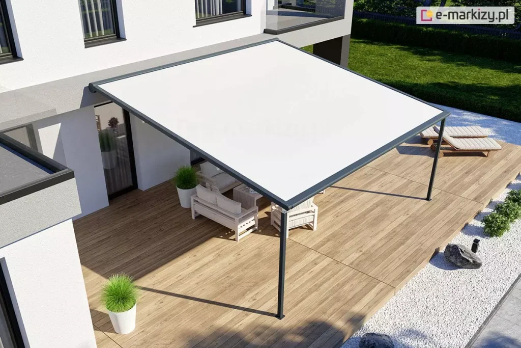 Widok z góry na nowoczesną pergolę tarasową V-Ziiip Aluprof o lekkiej konstrukcji aluminiowej w kolorze antracytowym, zamontowaną do elewacji białego, piętrowego budynku o prostej bryle. Zadaszenie stanowi gładka, napięta tkanina w kolorze białym, która całkowicie przykrywa strefę wypoczynkową na tarasie wykonanym z jasnobrązowych desek kompozytowych. Pod pergolą znajduje się zestaw mebli ogrodowych z jasnego drewna z białymi poduszkami oraz białe donice z dekoracyjną, zieloną trawą. W bocznej części tarasu, poza obrysem zadaszenia, ustawione są dwa drewniane leżaki basenowe. Teren wokół tarasu został starannie zaaranżowany – bezpośrednio przy deskach znajduje się pas białego żwiru dekoracyjnego z ozdobnymi kamieniami i niskimi nasadzeniami roślinnymi, a dalej rozciąga się zadbany, zielony trawnik ogrodzony niskim żywopłotem. Zdjęcie wykonane w pełnym świetle dziennym, prezentuje minimalistyczny i elegancki design systemów zadaszeń w nowoczesnym budownictwie jednorodzinnym.