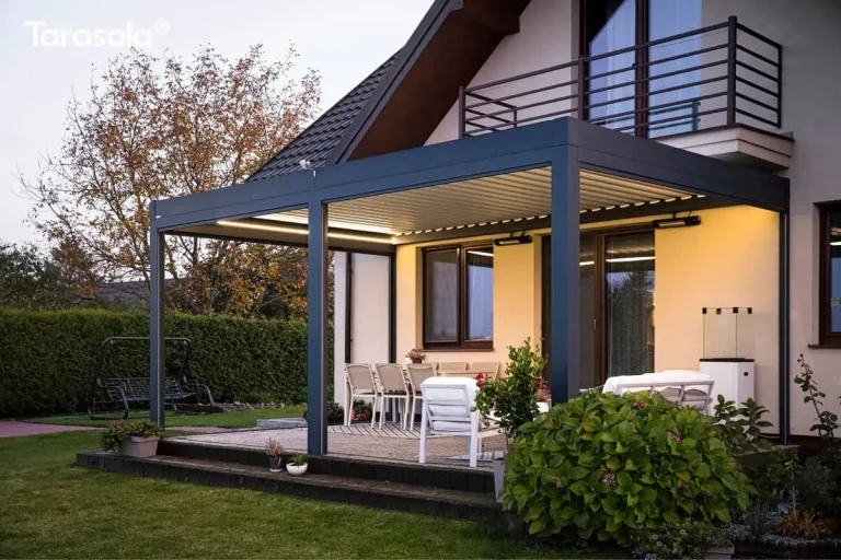 Zdjęcie przedstawia nowoczesną pergolę Essential Tarasola w porze zmierzchu, co pozwala na zaprezentowanie pełnego systemu zintegrowanego oświetlenia. Dwumodułowa konstrukcja w kolorze ciemnoantracytowym, zamontowana do jasnej elewacji domu, jest efektownie rozświetlona dzięki liniowym listwom LED zamontowanym wzdłuż wewnętrznych krawędzi ramy dachowej. Światło o ciepłej barwie odbija się od białych, zamkniętych lameli dachu oraz oświetla przestrzeń tarasu, na którym znajduje się zestaw mebli jadalnianych i wypoczynkowych. Na ścianie budynku, pod dachem pergoli Essential Tarasola, widoczne są dwa promienniki ciepła, podkreślając funkcjonalność tarasu jako salonu zewnętrznego dostępnego również w chłodniejsze wieczory. Po prawej stronie, obok białych foteli, stoi podświetlony, szklany ogrzewacz gazowy, stanowiący dodatkowe źródło światła i ciepła. Cała strefa wypoczynkowa, wyłożona jasną kostką brukową i wykończona ciemną palisadą, kontrastuje z pogrążonym w mroku ogrodem. W tle po lewej stronie widoczny jest zarys huśtawki ogrodowej na trawniku oraz ciemna ściana żywopłotu, natomiast na pierwszym planie po prawej stronie dominują gęste, ciemnozielone liście krzewów. Architektura domu z podświetlonymi wnętrzami widocznymi przez okna oraz ciemnym, skośnym dachem tworzy spójne i luksusowe tło dla prezentowanej realizacji Essential Tarasola.